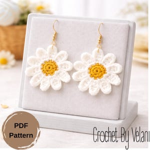May include: A pair of daisy-shaped crochet earrings with white petals and a yellow center, displayed on a gray stand. The earrings have gold-colored hooks. The image also includes the text "PDF Pattern" and "Crochet By Velani".