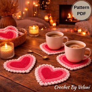 May include: Heart-shaped crochet coasters in shades of red and pink with white trim. Two coffee cups and a gold spoon sit on the coasters. The image also includes lit candles and a basket of coasters. Text reads "Pattern PDF" and "Crochet By Velani."