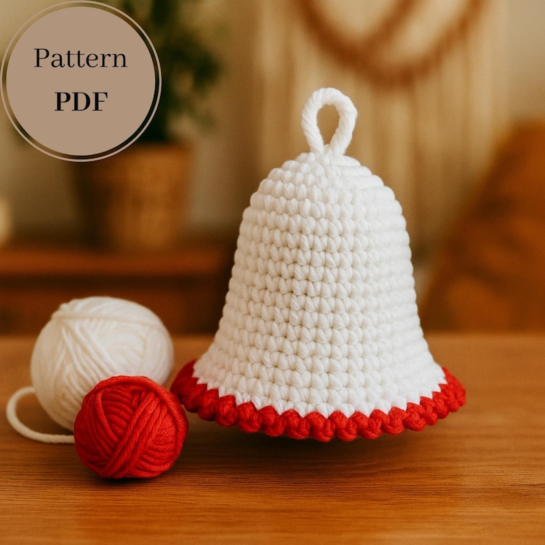 Op de afbeelding: Een witte gehaakte bel met een geschulpte rode rand en een lus aan de bovenkant. Een bol wit garen en een bol rood garen liggen naast de bel. De tekst "Pattern PDF" staat in een cirkel linksboven.
