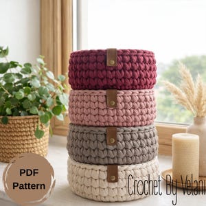 May include: A stack of four handmade crochet baskets in varying colors: burgundy, dusty rose, gray, and cream. Each basket has a leather strap closure. The image also includes a plant, a candle, and the text "PDF Pattern" and "Crochet By Velani".