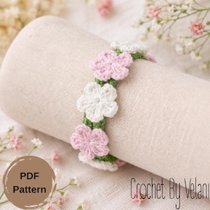 May include: A crochet bracelet featuring pink and white flower designs on a green band. The bracelet is displayed on a beige fabric roll. The image also includes the text "PDF Pattern" and "Crochet By Velani".