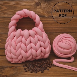 Op de afbeelding: Een lichtroze, handgebreide tas met een gevlochten handvat en een bijpassende bol garen. De tas is gemaakt van dik, zacht materiaal en ligt naast een hoop koffiebonen. De woorden "PATTERN PDF" staan in een cirkel rechtsboven.
