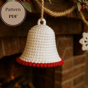 Op de afbeelding: Een witte gehaakte bel met een rode rand, hangend aan een touw. De bel hangt voor een bakstenen open haard. De tekst "Pattern PDF" staat in een cirkel linksboven.