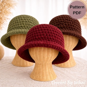 以下が含まれることがあります： オリーブグリーン、バーガンディ、ブラウンの3つのかぎ針編みのバケットハットが、柳のマネキンの頭に展示されています。帽子はテクスチャードステッチと小さなつばがあります。画像には「Pattern PDF」グラフィックと「Crochet By Velani」のテキストが含まれています。