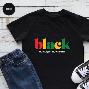 May include: Black t-shirt with the word "black" in red, yellow, and green, with the phrase "no sugar. no cream." printed below. The shirt is paired with blue jeans and black and white sneakers.
