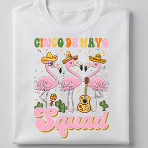 May include: White t-shirt featuring a Cinco de Mayo design. The graphic includes three pink flamingos wearing sombreros, a guitar, maracas, and the words "Cinco de Mayo Squad" in colorful text.