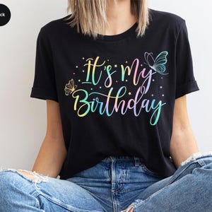 May include: Black t-shirt with the text "It's my Birthday" in a colorful, pastel font. The shirt features butterfly graphics and small star accents. The sleeves are cuffed.