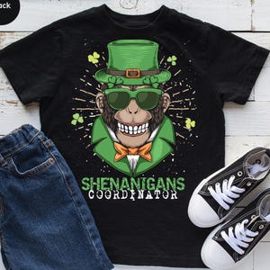 May include: Black t-shirt featuring a cartoon chimpanzee wearing a green top hat, sunglasses, and bow tie. The text "SHENANIGANS COORDINATOR" is printed below the graphic. A pair of black and white sneakers and blue jeans are also visible.