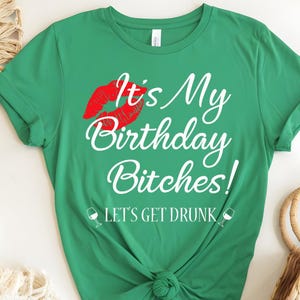 May include: A green t-shirt with the text "It's My Birthday Bitches!" in white script, a red lip graphic, and the phrase "LET'S GET DRUNK" with wine glass icons. The shirt is made of a soft material.