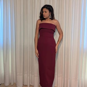 May include: A burgundy strapless dress with a fitted silhouette. The dress features a folded neckline and falls to the ankles. The model is wearing gold-colored heeled sandals and gold jewelry. The background is a white curtain.