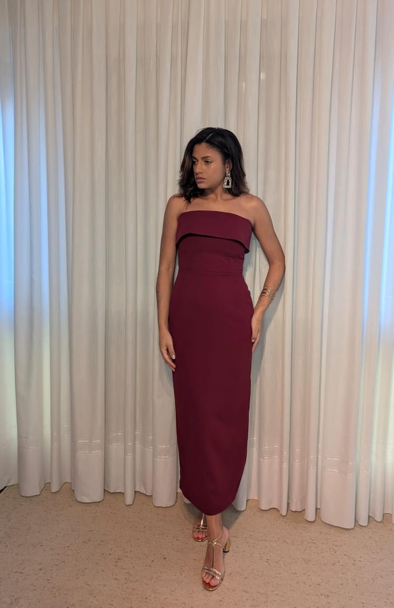 May include: A burgundy strapless dress with a fitted silhouette. The dress features a folded neckline and falls to the ankles. The model is wearing gold-colored heeled sandals and gold jewelry. The background is a white curtain.