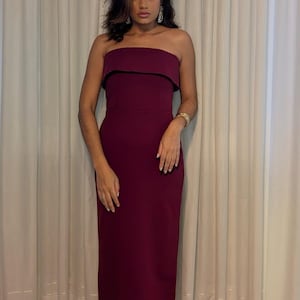 May include: A strapless, floor-length burgundy dress with a black trim. The dress is worn with gold sandals and a gold bracelet. The background features white curtains and a light-colored floor.