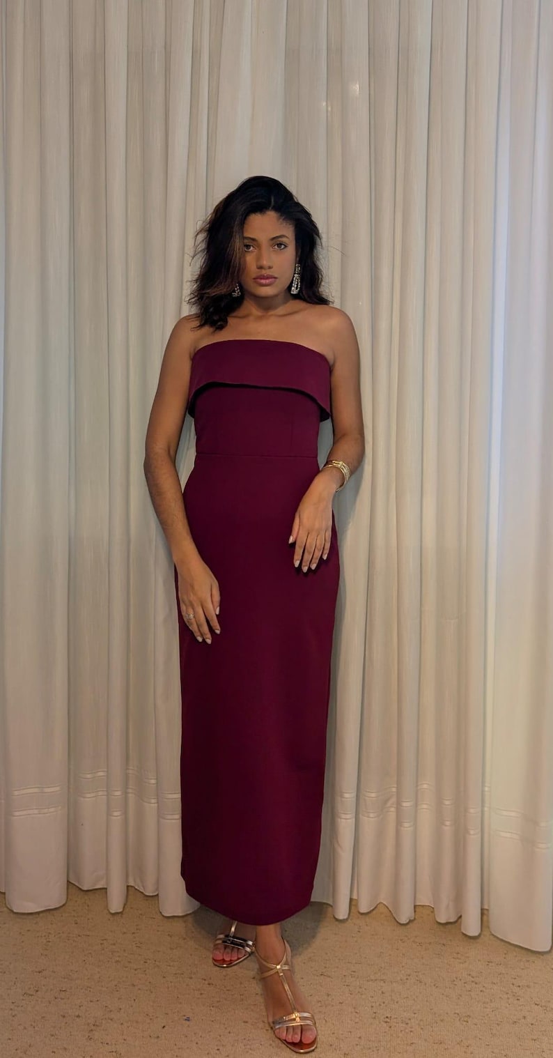 May include: A strapless, floor-length burgundy dress with a black trim. The dress is worn with gold sandals and a gold bracelet. The background features white curtains and a light-colored floor.