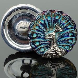 May include: Two decorative buttons. One is a shiny silver metal button. The other is a round button with a peacock design. The peacock has a silver body and a fan of iridescent blue and teal feathers.