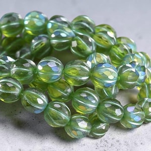 May include: A collection of round, translucent green glass beads featuring a faceted, melon-like texture. Each bead displays a subtle iridescent finish that reflects hints of blue and gold light across the surface of the glass material.