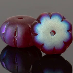 Op de afbeelding: Twee iriserende glazen kralen. De kralen hebben een dieprode-paarse kleur met een bloemachtige vorm. Elke kraal heeft een centraal gat en geëtste lijnen. De randen van de kralen hebben een blauwe en groene glans.