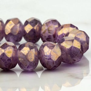 Puede incluir: Primer plano de cuentas moradas facetadas con detalles dorados. Las cuentas tienen un diseño geométrico y un brillo metálico. Las cuentas están dispuestas en una fila, lo que sugiere que son para la fabricación de joyas o manualidades.