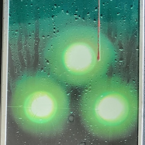 Op de afbeelding: Een rechthoekig, metalen object met een groene en zwarte achtergrond. Drie heldergroene cirkels zijn zichtbaar, met een rode streep die verticaal loopt. Het oppervlak heeft een natte, druppelachtige textuur.