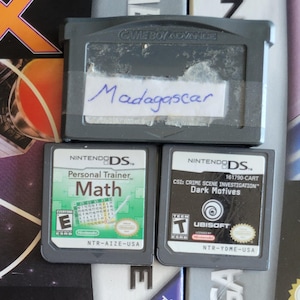 Puede incluir: Una colección de cartuchos de juegos de Nintendo DS y Game Boy Advance. Los títulos incluyen "Madagascar", "Personal Trainer: Math" y "CSI: Crime Scene Investigation: Dark Motives". Los cartuchos son grises y negros con ilustraciones coloridas.