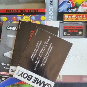 Op de afbeelding: Een verzameling Game Boy- en Classic NES Series-gamecartridges en handleidingen. De afbeelding toont een Dr. Mario-cartridge, een Pac-Man-gamebox en diverse instructieboekjes. Het Game Boy-logo is zichtbaar op de handleidingen.