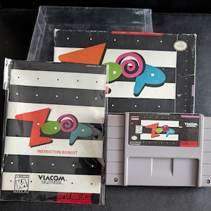 Puede incluir: Un conjunto de cartucho de videojuego Super Nintendo, folleto de instrucciones y caja. El título del juego "Zoop" se muestra en rojo, verde, azul y rosa. El folleto de instrucciones tiene un fondo a rayas blanco y negro. La caja tiene un triángulo naranja.