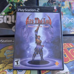 Op de afbeelding: PlayStation 2 game case voor "Arc The Lad: Twilight of the Spirits". De cover toont een mannelijk personage met een opgeheven arm, tegen een paarse en blauwe achtergrond. De case wordt omringd door andere game cases.