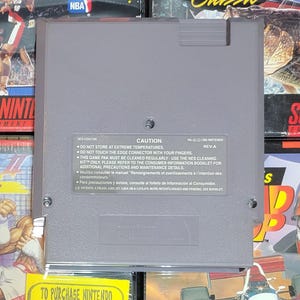 K&ouml;nnte beinhalten: Eine graue Nintendo NES-Spielkassette. Die R&uuml;ckseite der Kassette hat ein Vorsichtsetikett mit Text in Englisch, Franz&ouml;sisch und Spanisch. Das Etikett enth&auml;lt Warnungen bez&uuml;glich Temperatur und Reinigung.