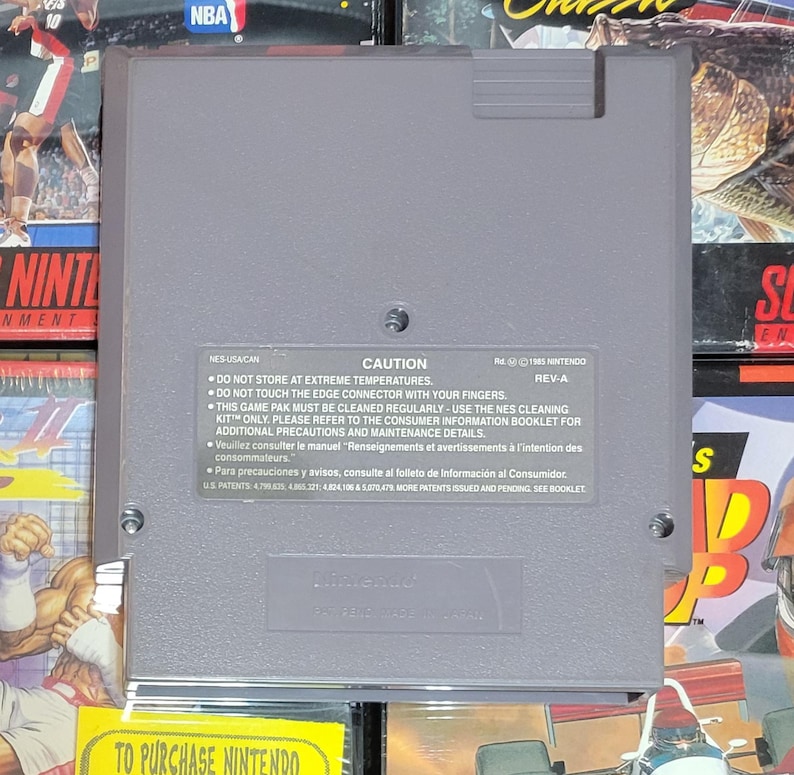 K&ouml;nnte beinhalten: Eine graue Nintendo NES-Spielkassette. Die R&uuml;ckseite der Kassette hat ein Vorsichtsetikett mit Text in Englisch, Franz&ouml;sisch und Spanisch. Das Etikett enth&auml;lt Warnungen bez&uuml;glich Temperatur und Reinigung.