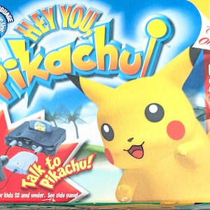 Puede incluir: Caja del juego de Nintendo 64 "Hey You, Pikachu!" con un gran personaje amarillo de Pikachu. La caja incluye el paquete de juego, la unidad de reconocimiento de voz y el micrófono. También es visible el texto "Talk to Pikachu!".