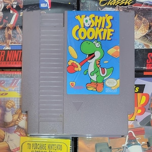 K&ouml;nnte beinhalten: Eine graue Nintendo-Spielkassette mit dem Spiel "Yoshi's Cookie." Das blaue Etikett zeigt den Spieletitel und eine Yoshi-Cartoonfigur. Andere Spielboxen umgeben die Kassette.