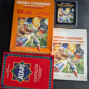 May include: A vintage Atari Missile Command video game set including an orange cardboard box, a black plastic game cartridge, a paper instruction manual, and a red Atari catalogue, all featuring retro space-themed illustrations and text.
