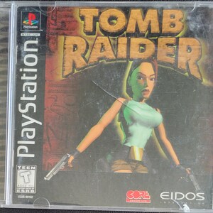 Puede incluir: Caja de juego de PlayStation para Tomb Raider. La portada presenta a un personaje femenino con dos pistolas, con el título del juego en letras grandes y estilizadas. La caja está en una funda de plástico transparente.
