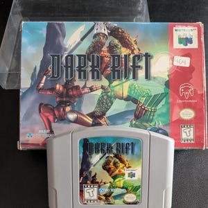 Puede incluir: Un cartucho y caja de videojuego de Nintendo 64 para "Dark Rift". La carátula muestra una escena de fantasía con dos figuras luchando. El cartucho es gris con el título del juego y un gráfico similar.