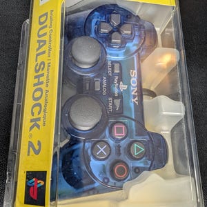 Può includere: Un controller analogico DualShock 2 blu nella sua confezione originale. Il controller ha pulsanti neri e pad direzionali. La confezione è gialla e presenta il logo PlayStation.