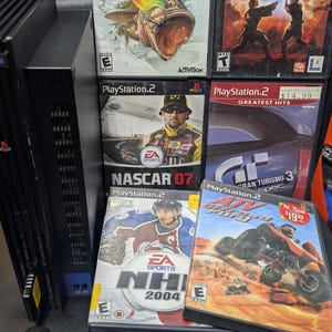 May include: A black PlayStation 2 console with several game cases, including "Rapala Pro Fishing," "Star Wars Episode III," "NASCAR 07," "NHL 2004," and "ATV Offroad Fury." Also included are several black Sony controllers.