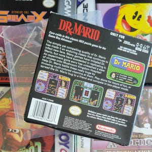 Op de afbeelding: Een Dr. Mario Game Boy Advance videospeldoos. De zwarte doos heeft kleurrijke graphics en tekst, waaronder de speltitel en screenshots. Het zit in een beschermende plastic hoes, met andere speldozen op de achtergrond.