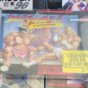Puede incluir: Caja sellada de cartucho de juego Super Nintendo Entertainment System para Street Fighter II Turbo. La ilustración de la caja presenta personajes musculosos en poses de lucha. Una etiqueta amarilla dice: "TO PURCHASE NINTENDO CARTRIDGE, BRING THIS BOX TO FRONT COUNTER."