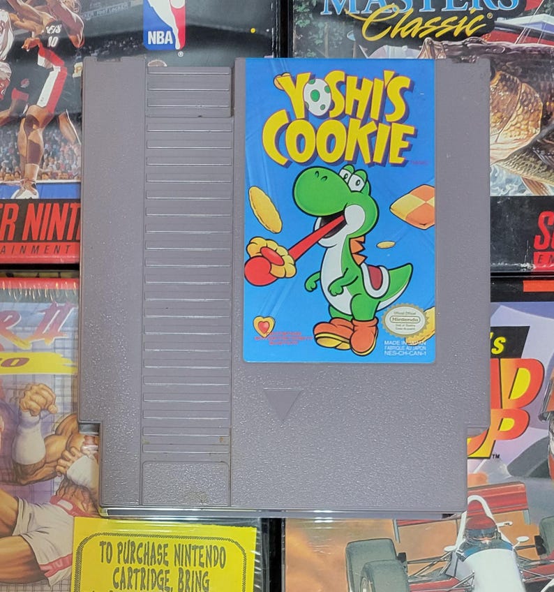 K&ouml;nnte beinhalten: Eine graue Nintendo-Spielkassette mit dem Spiel "Yoshi's Cookie". Das Etikett zeigt Yoshi, einen gr&uuml;nen Dinosaurier, mit roter Zunge und einem Keks. Der Hintergrund ist blau mit gelben Keksen. Andere Spielboxen umgeben die Kassette.