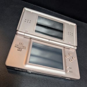 May include: A silver Nintendo DS handheld game console, open and displayed on a black surface. The console features two screens, a directional pad, and action buttons. The device is in good condition.