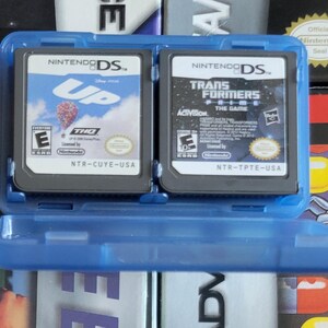 Puede incluir: Una funda azul para juegos de Nintendo DS con dos cartuchos de juego. Uno muestra el logotipo de la película "UP", y el otro el logotipo del juego "Transformers". La funda está abierta, mostrando los cartuchos.