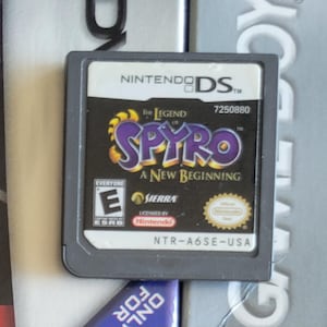 Puede incluir: Un cartucho de juego de Nintendo DS para "The Legend of Spyro: A New Beginning". El cartucho es negro con una etiqueta colorida con el título del juego en morado y amarillo. La clasificación ESRB es "E" para Todos.