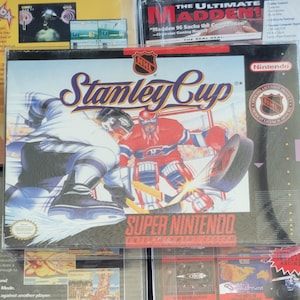 May include: A Super Nintendo video game box for "Stanley Cup" featuring hockey players in action. The box art shows a player in white and blue skating towards a goalie in red. The Nintendo logo and NHL emblem are visible.