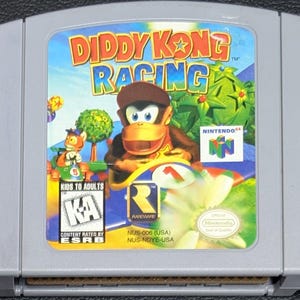 May include: A gray Nintendo 64 video game cartridge for Diddy Kong Racing. The label features Diddy Kong in a race car, the game title in yellow and blue text, the Nintendo 64 logo, a Rareware logo, and an ESRB Kids to Adults rating symbol.