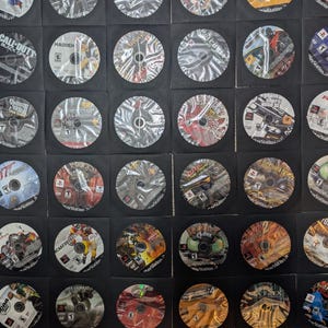 Puede incluir: Una colección de discos de juegos de PlayStation, cada uno en una funda de papel negro. Los discos presentan arte y texto coloridos, incluyendo títulos como "Call of Duty", "Madden" y "Lord of the Rings". La disposición es en forma de cuadrícula.