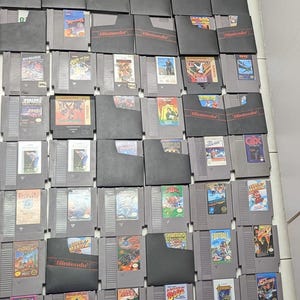 Peut inclure: Une grande collection de cartouches de jeu rectangulaires grises pour Nintendo Entertainment System disposées en grille. Chaque cartouche présente une étiquette colorée unique avec divers titres et illustrations, certaines étant noires.