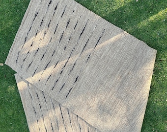 Puede incluir: Una alfombra rectangular tejida de color beige con detalles de rayas verticales negras. La alfombra está doblada, revelando un detalle de flecos en el borde inferior. La alfombra está hecha de fibra natural y se muestra sobre una superficie de hierba verde.