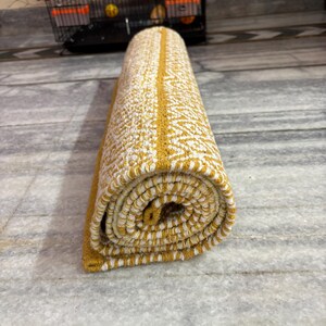 Puede incluir: Alfombra enrollada con un patr&oacute;n amarillo y blanco. La alfombra tiene una franja central amarilla y un dise&ntilde;o texturizado. La alfombra est&aacute; enrollada, mostrando el patr&oacute;n en los bordes. Est&aacute; sobre un suelo de m&aacute;rmol.