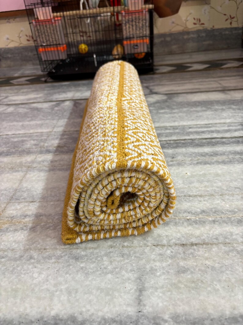 Puede incluir: Alfombra enrollada con un patr&oacute;n amarillo y blanco. La alfombra tiene una franja central amarilla y un dise&ntilde;o texturizado. La alfombra est&aacute; enrollada, mostrando el patr&oacute;n en los bordes. Est&aacute; sobre un suelo de m&aacute;rmol.