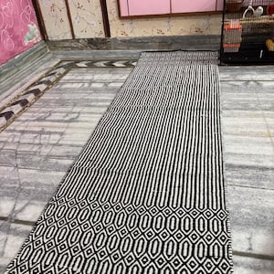 May include: A long, rectangular black and white rug with a geometric pattern. The rug features vertical stripes in the center and a detailed diamond and oval design along the border. The rug is made of woven material.