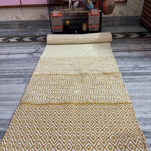 Puede incluir: Una alfombra rectangular enrollada con un patr&oacute;n geom&eacute;trico. La alfombra presenta bandas alternas de crema y amarillo mostaza, con un patr&oacute;n de diamantes en la secci&oacute;n inferior. La alfombra est&aacute; hecha de material tejido.